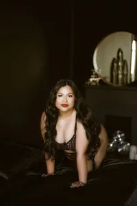 A confident woman with long dark hair kneels on a black bed in a dim, elegant room, gazing at the camera; warm light creates a moody tone.