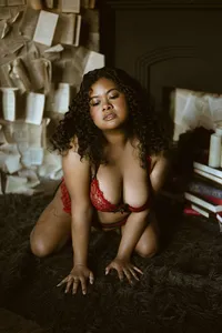 Adult woman with curly hair kneels on a dark rug in red lace lingerie, surrounded by books and papers, gazing calmly toward the camera.
