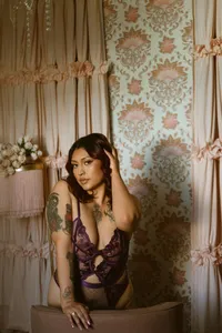 Woman with tattoos in purple lace lingerie poses behind a chair in a warm vintage room with floral wallpaper, gathered curtains, and soft light.