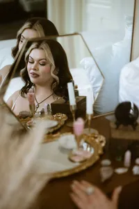 Woman with wavy hair applies makeup at a vanity, reflected in a mirror among cosmetics and jewelry, creating an intimate, serene mood.