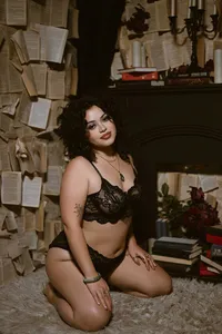 Curly-haired woman kneels in black lace lingerie in a vintage study, surrounded by books and papers, creating a moody, intimate portrait.