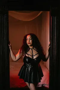 Curly-haired woman in a black leather dress poses in a doorway, hands raised, against a red-lit bedroom backdrop; confident, sultry mood.