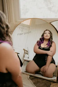 Plus-size woman with purple-tipped hair poses confidently in black lingerie before a vintage mirror, with “PINK DOOR” reflected behind her.