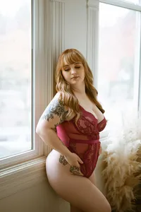 Curvy red-haired woman in red lace lingerie poses by a sunlit window, showing floral tattoos and a serene, confident expression.