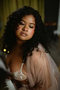 Portrait of a woman with voluminous curly hair in a sheer blush robe and lace top, gazing thoughtfully in warm, moody indoor light.