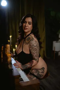 Tattooed woman in a black outfit plays piano in a dim, candlelit room, gazing down with a calm, sultry expression.