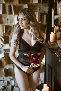 Woman with long wavy hair in black lace holds red roses in a candlelit vintage room, gazing aside with a thoughtful, romantic expression.