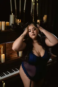 Curvy woman in a blue dress poses beside a piano, hands in her hair, surrounded by glowing candles in a dark, intimate, dramatic room.
