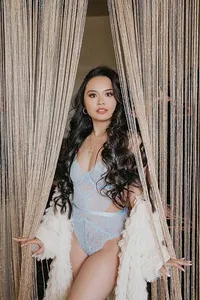 Confident woman with long dark curls parts beaded curtains, wearing a pale blue bodysuit and white feathered coat in a glamorous portrait.