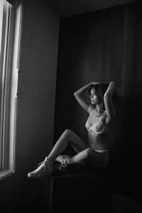 Black-and-white portrait of a woman in lingerie and sneakers seated by a tall window, arms raised, creating a moody, dramatic atmosphere.