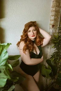 Red-haired woman in a dark green outfit poses among lush houseplants beside a rustic stone wall, gazing confidently at the camera.