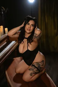 Woman with dark hair and tattoos kneels beside a piano in black attire, lit by candles and a spotlight, creating an intimate, sultry mood.