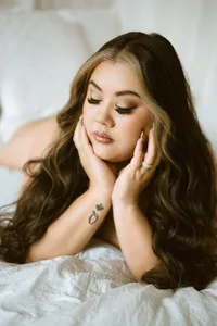Portrait of a woman with long, wavy brown hair resting on white bedding, eyes closed and hands cupping her face; soft light creates a serene mood.