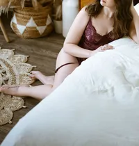 Woman in burgundy lace lingerie reclines beside a white bed, bare legs on a woven rug; a warm, rustic bedroom creates a calm, intimate mood.