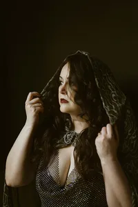 Portrait of a woman lifting a dark patterned veil, her long hair framing a jeweled chainmail dress in a dramatic, mysterious mood.