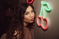 Portrait of a curly-haired woman blowing a kiss beside a glowing neon cherry sign, creating a playful, warm, retro nightlife mood.