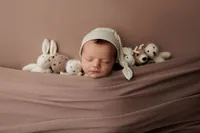 newborn baby wearing cream colored hat in brown wrap surrounded by little stuffed animals during luxury studio session at CG Photography Los Angeles