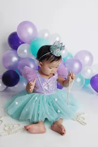 baby with tiara in adorable turquoise and purple outfit with purple, white, and turquoise balloons in background during studio milestone photography session in North Hollywood