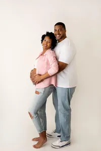 smiling expecting couple standing while holding baby bump in studio portrait for maternity photoshoot in North Hollywood