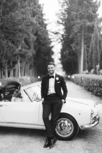 Groom in tuxedo leaning on vintage Italian car – stylish portrait at luxury Italy wedding.