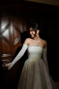 Bride in a high-fashion, off-the-shoulder lace gown with long gloves, featuring a polished Kohnur elegant bridal updo and flawless timeless wedding makeup, posing in a dramatic, dimly lit setting.
