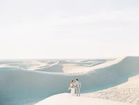 Kohnur epic couple portrait with bride in natural minimal makeup and sleek low updo wearing a flowy gown on vast white sand dunes in Glamis, California