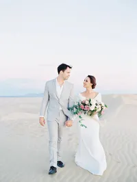 Kohnur romantic couple portrait with bride in natural subtle makeup and a sleek low bun walking on soft sand dunes at sunset in Glamis, California