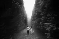 Groom holding the brides dress as they walk between some trees