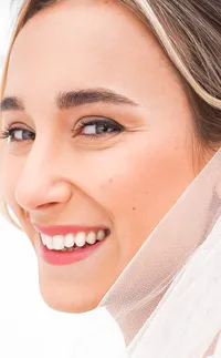 A bride grins happily into the camera. Her beauty is radiant and soft. She wears a peachy pink lip for her wedding day.