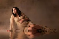 Pregnant woman in a soft, draped white gown cradling her belly while seated on a floor in a bright Waukesha studio, showcasing wardrobe options provided by the photographer.