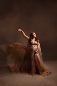 Pregnant woman wearing a flowing mocha tulle gown with embroidered bodice, captured mid-movement with a dramatic painterly backdrop in a Brookfield-area photography studio.