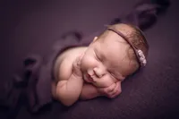 Sleeping baby girl posed with hands under chin on a purple blanket, wearing a matching lavender headband during a newborn session near Lake Country, WI.