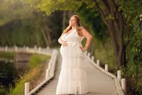 Pregnant woman in a tiered white gown standing confidently on the Delafield boardwalk, surrounded by lush green trees and soft sunset light during a serene outdoor maternity session.