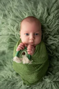 Newborn baby with bright blue eyes swaddled in green, holding a tiny rainbow toy on a fluffy green blanket at a Waukesha, Wisconsin photography studio.