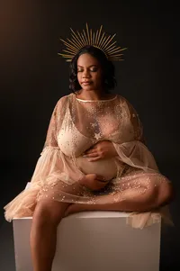 Pregnant Black woman in gorgeous shimmery gold bodysuit and crown, sitting upon a stool.