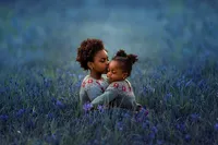Two Black sisters share an embrace in a field of blue wildflowers.