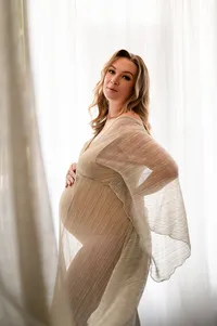 Pregnant woman standing in front of sheer white curtains, softly draped in a translucent gold-toned gown, glowing in natural window light during a fine art maternity session in Waukesha, WI.