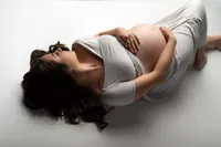 Pregnant woman in a soft gray gown lying on a seamless white studio floor, gently holding her belly with eyes closed during a peaceful, minimalist maternity session in Waukesha near Hartland, Wisconsin.