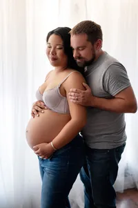 Pregnant woman in a lavender bra and jeans standing in soft window light, with her partner embracing her from behind during a cozy, love-filled maternity session in Waukesha, Wisconsin.