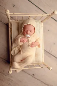 Cozy baby photo of a newborn in a cream knit romper and bonnet, sleeping in a vintage bed at a Brookfield-area newborn photo studio.