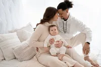 Indoor studio portrait of a mother, father and baby boy dressed all in white against backlit sheer curtains at our Waukesha location.