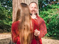 Man secretly holds an engagement ring behind his back and makes a shh gesture while standing with his girlfriend during a proposal photo session in a Los Angeles park.