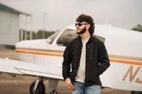 Senior portrait at a small hangar near Mobile International Airport in Mobile, Alabama — black
jacket and aviators in front of a small airplane, looking to the side.