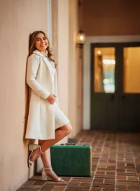 Senior portrait at the Malaga Inn in Mobile, Alabama— wearing a vintage white coat and dress leaning against a while stand next to a green suitcase.