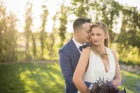 A groom in a navy suit holds his bride close in golden evening light, her plum bouquet in her hands.