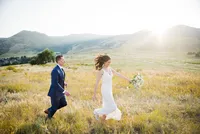 A bride and groom walk hand in hand through tall grass with the Colorado foothills behind them.
