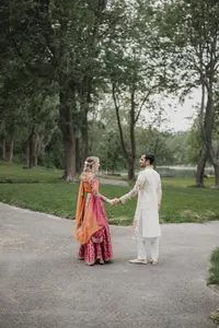 Couple se tenant la main sur un sentier, mariée en lehenga orange et rose éclatant, marié en kurta blanc brodé, entourés de verdure lors de leur cérémonie du Mehndi