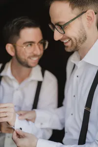 Mariés riant ensemble pendant les préparatifs, chemises blanches et bretelles, lunettes et alliance visibles, moment de complicité avant la cérémonie