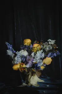 Bouquet de mariage composé de renoncules jaunes, delphiniums bleus et roses pâles, éclairage dramatique sur fond de velours sombre, style pictural inspiré des maîtres hollandais
