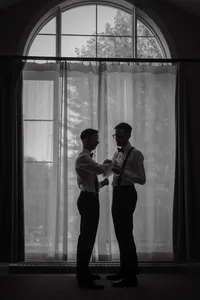Moment intime et complice entre deux mariés LGBTQ lors des préparatifs, devant une grande fenêtre voilée dans les Laurentides — une scène élégante et authentique, capturée par un photographe de mariage à Montréal, célébrant la diversité, l’amour et la tendresse de leur connexion.